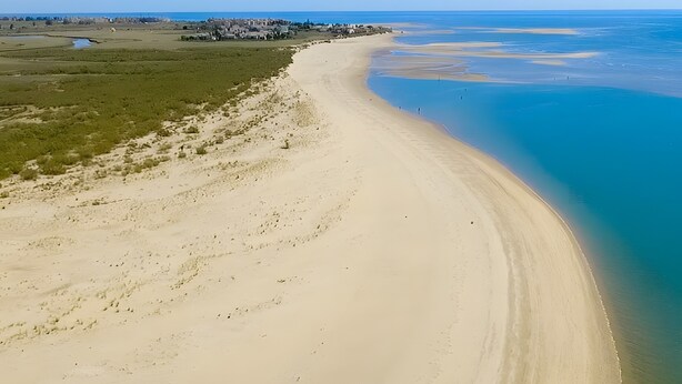 Un paysage côtier avec une plage de sable, une eau d’un bleu limpide et une agglomération visible au loin.