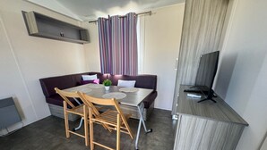Un espace de vie compact comprenant une table à manger, deux chaises, un canapé, un téléviseur posé sur un meuble et une fenêtre avec des rideaux.