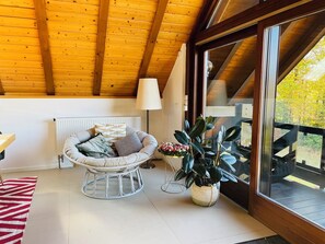 Une pièce chaleureuse avec un plafond en bois, un radiateur blanc, un fauteuil rembourré, une plante en pot et une vue à travers une porte vitrée.