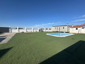 A backyard with artificial grass, a swimming pool, a small house, and a fenced area.