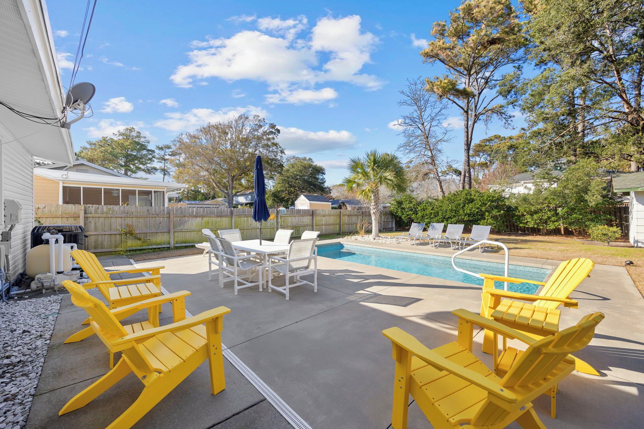 A backyard with a swimming pool, yellow Adirondack chairs, a dining table, and a blue umbrella.