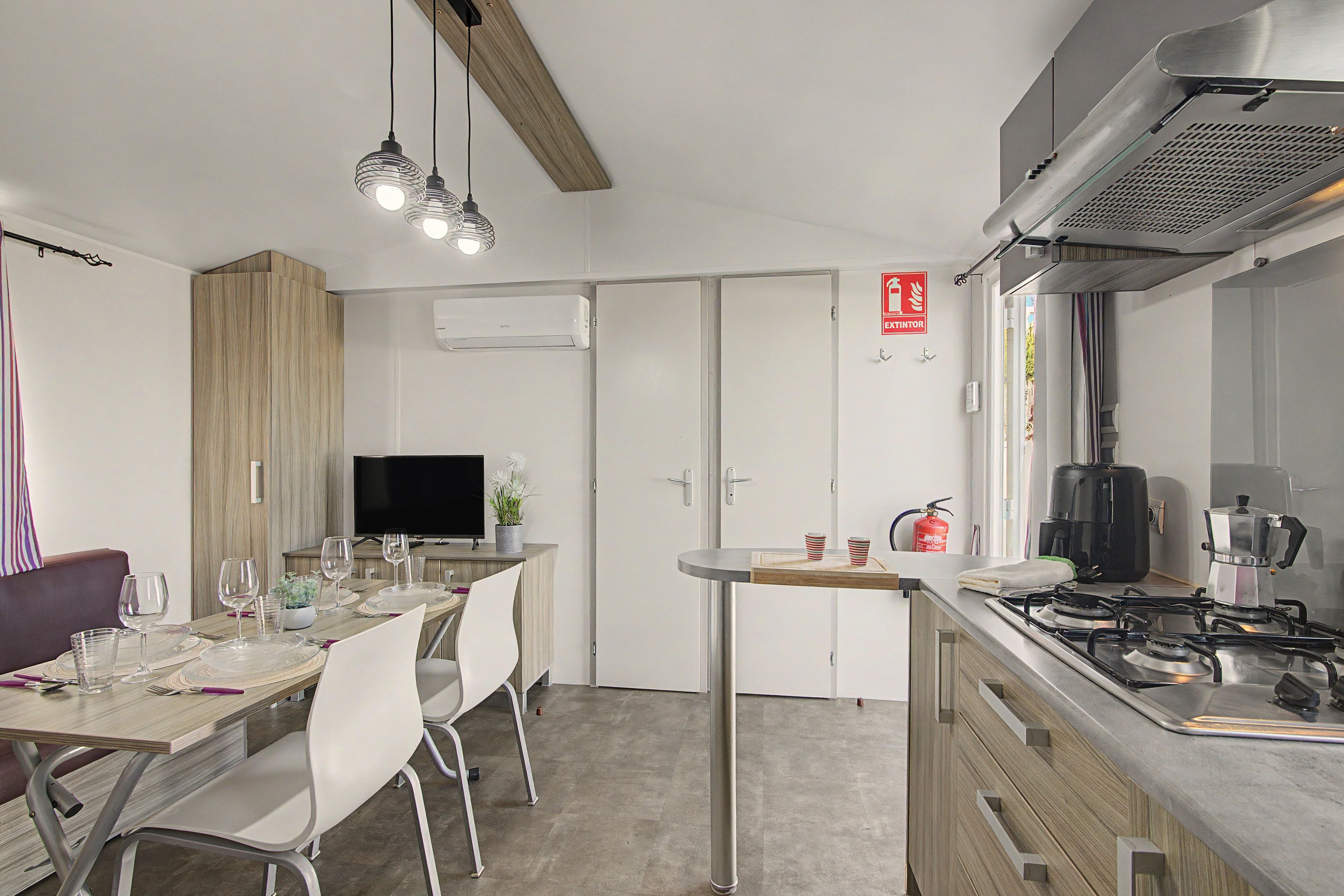 Una cocina moderna con estufa a gas, cafetera y zona de comedor con televisor y planta en maceta.