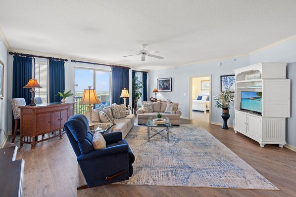 A spacious living room with a large sofa, a coffee table, and a television. There is a ceiling fan, a cabinet with decorative items, and a view of the outdoors through the windows. - Margate 2109 in Kingston, Oceanview, Pool, Beach (Myrtle Beach)