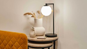 A round wooden table with a white vase containing dried plants, a black floor lamp, and a small book. - Host Stay Holmfirth Hideaway (Holmfirth)