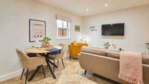 A modern living room with a sofa, a dining table with chairs, a TV mounted on the wall, and a window with a view. - Host Stay Holmfirth Hideaway (Holmfirth)