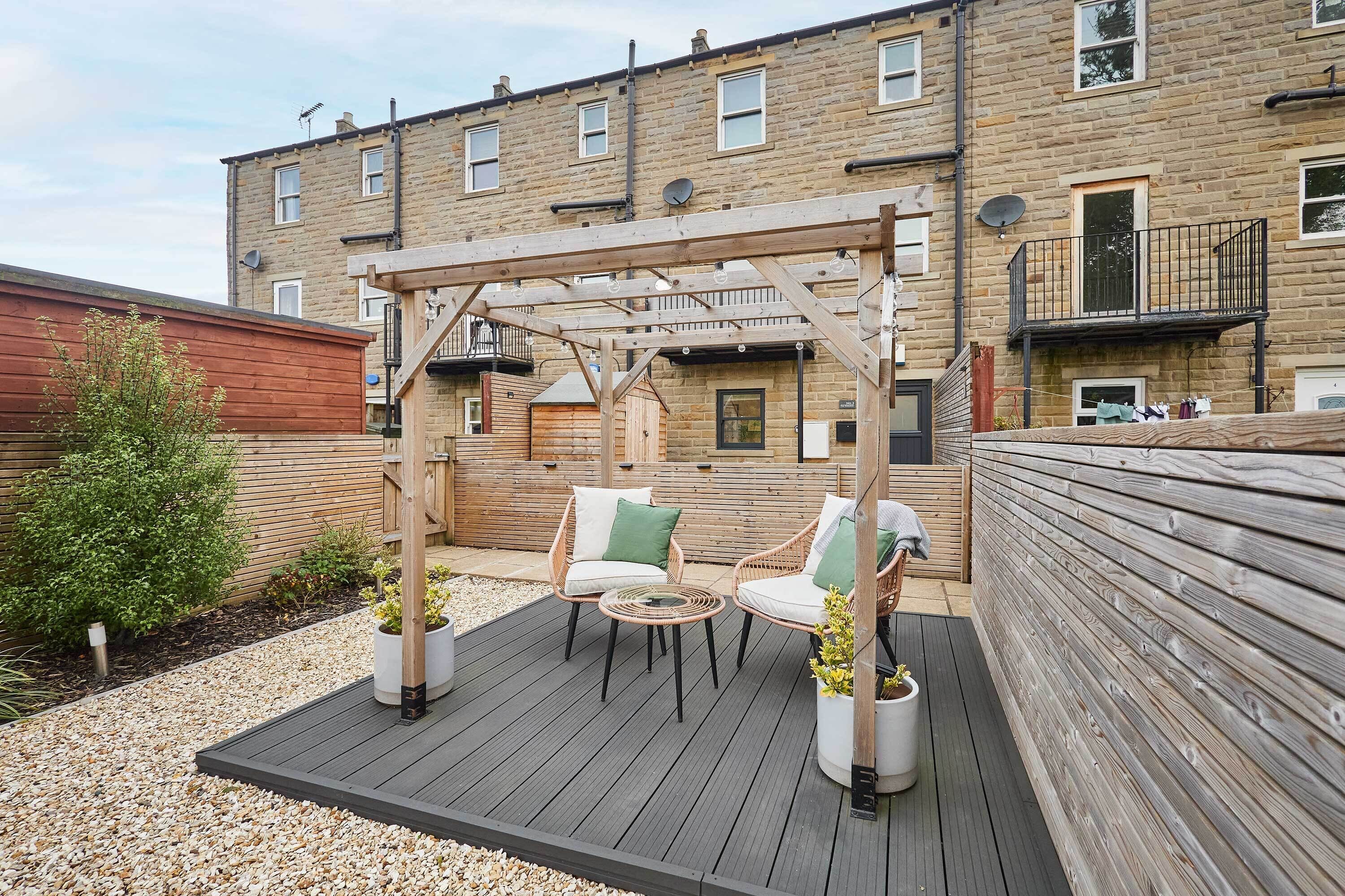Eine Holzterrasse mit Gartenmöbeln, einer Pergola und einem Backsteinhaus im Hintergrund.
