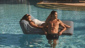 Un homme et une femme se détendent sur une plateforme flottante à rayures, dans une piscine à l’eau claire.