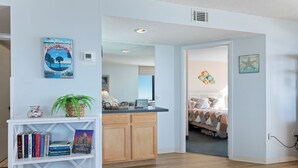 A modern living room with a white bookshelf, a small kitchen area, and a bedroom with a bed and decorative wall art. - Sands Beach Club 814, End Unit Sunsets & Pools (Myrtle Beach)