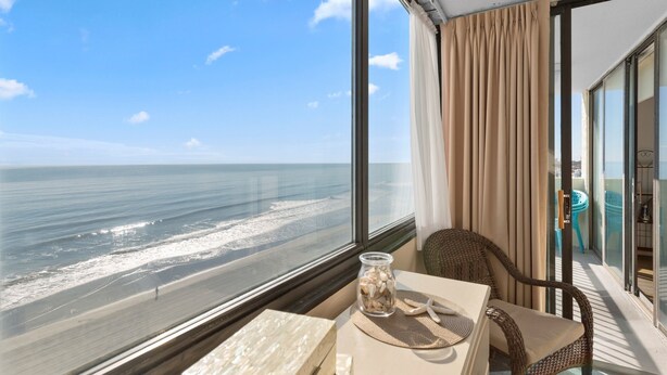 A room with a large window overlooking a beach, a wicker chair, a table with a vase of seashells, and a view of the ocean. - Sands Beach Club 814, End Unit Sunsets & Pools (Myrtle Beach)