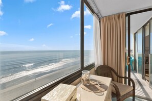 A room with a large window overlooking a beach, a wicker chair, a table with a vase of seashells, and a view of the ocean. - Sands Beach Club 814, End Unit Sunsets & Pools (Myrtle Beach)