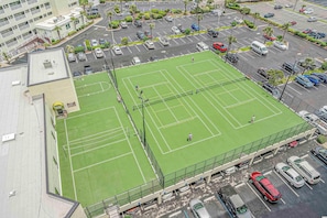 An aerial view of a tennis court surrounded by a parking lot with multiple cars and a building in the background.