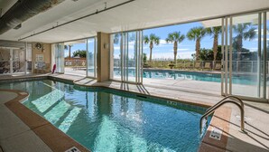 En inomhuspool med väggar av genomskinligt glas, utsikt över palmer och en skylt som visar var livräddaren finns.