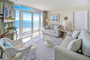 Een ruime woonkamer met een grote schuifpui naar een balkon met uitzicht op de oceaan. Er staat een witte bank, een rieten fauteuil en een glazen salontafel.
