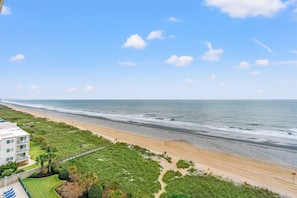 A beach view with a building in the foreground, a sandy path, and a clear sky.