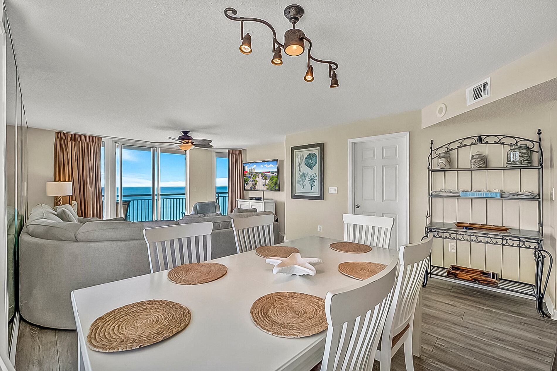 A dining area with a white table set with placemats, surrounded by white chairs. There is a ceiling fan above the table, and a kitchen area is visible in the background.