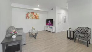 Un salon moderne avec un canapé gris, une table basse en verre, un fauteuil à motifs, un téléviseur posé sur un meuble blanc et un vase orné de motifs floraux.