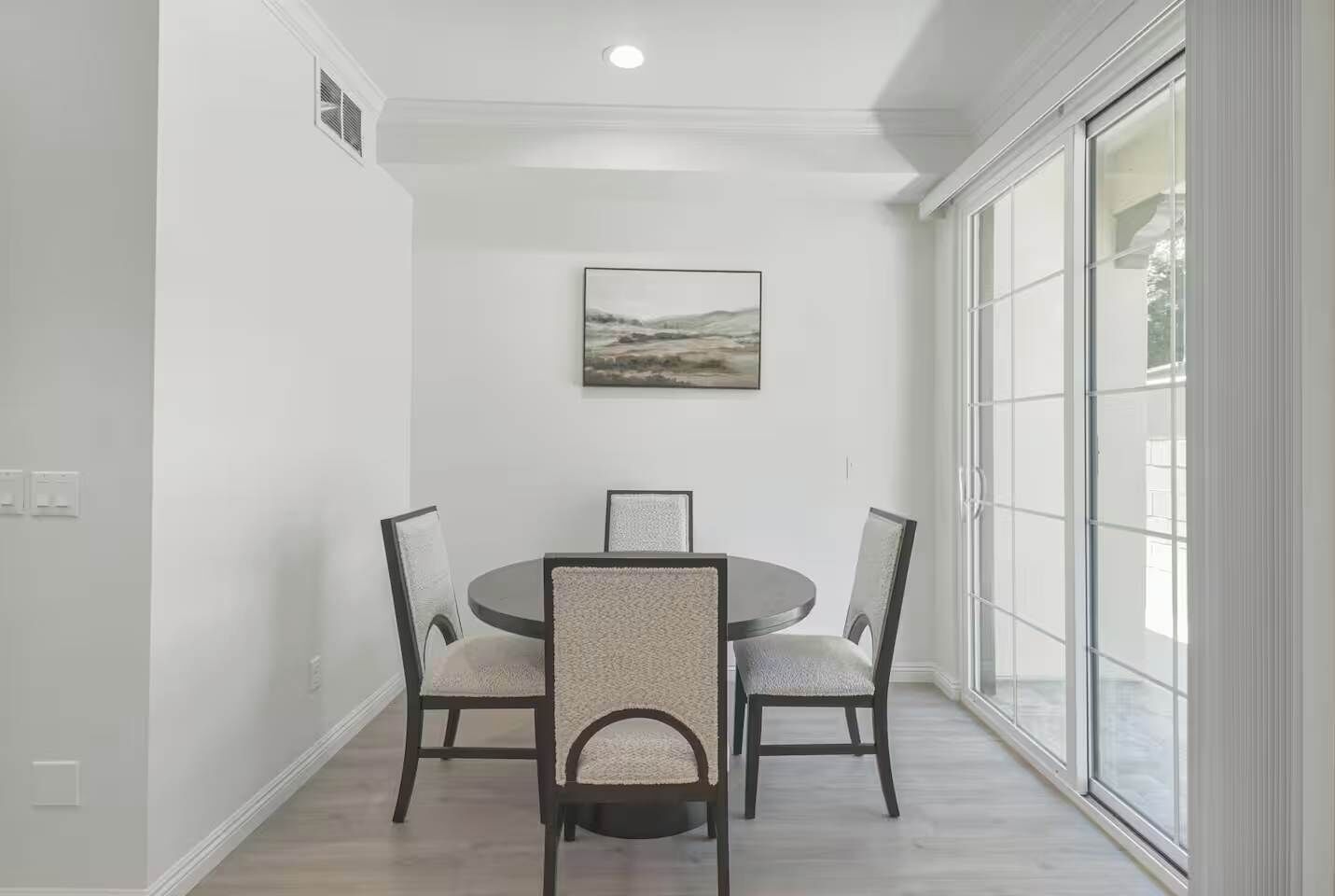 A dining area with a round table and four chairs, a painting on the wall, and a large window with a view.