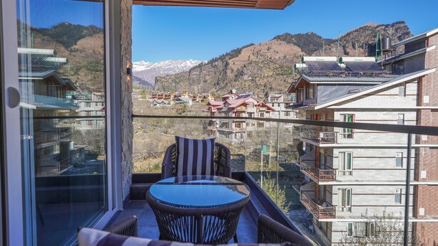 A balcony with a glass door, wicker furniture, and a view of a mountain town with buildings and snow-capped peaks. - Carom Hotel Manali (Manali)