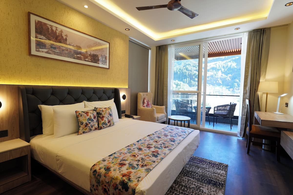 A hotel room with a large bed, a desk, a chair, and a balcony with a view of the mountains.