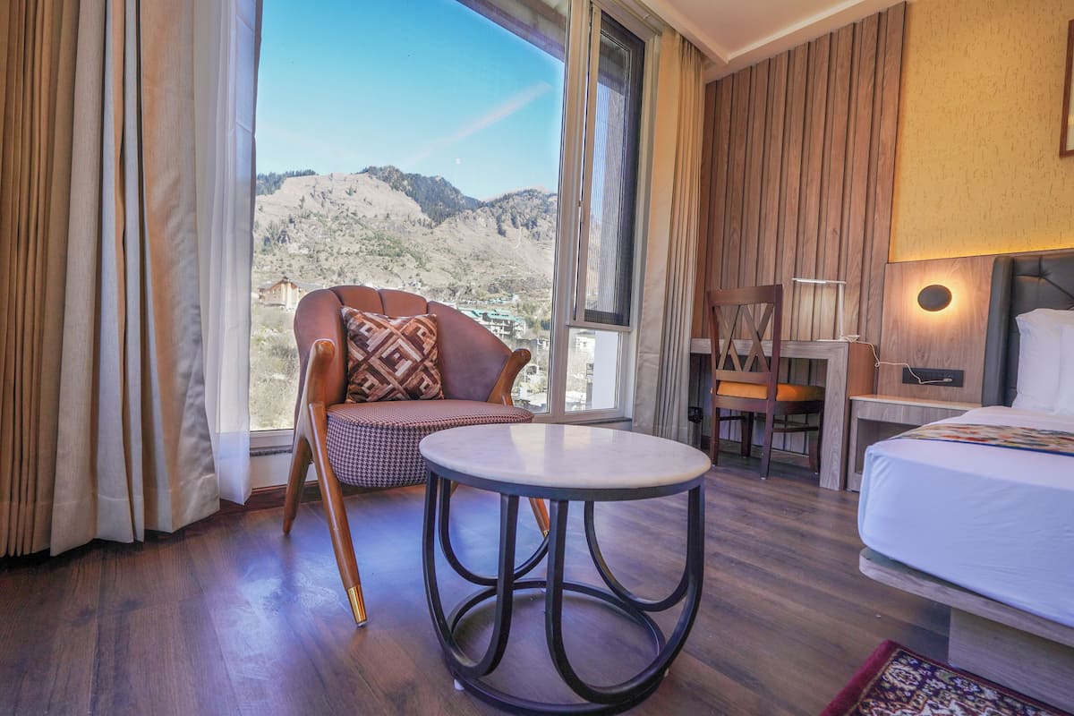 A hotel room with a large window, a round table, a bed, and a chair with a cushion.