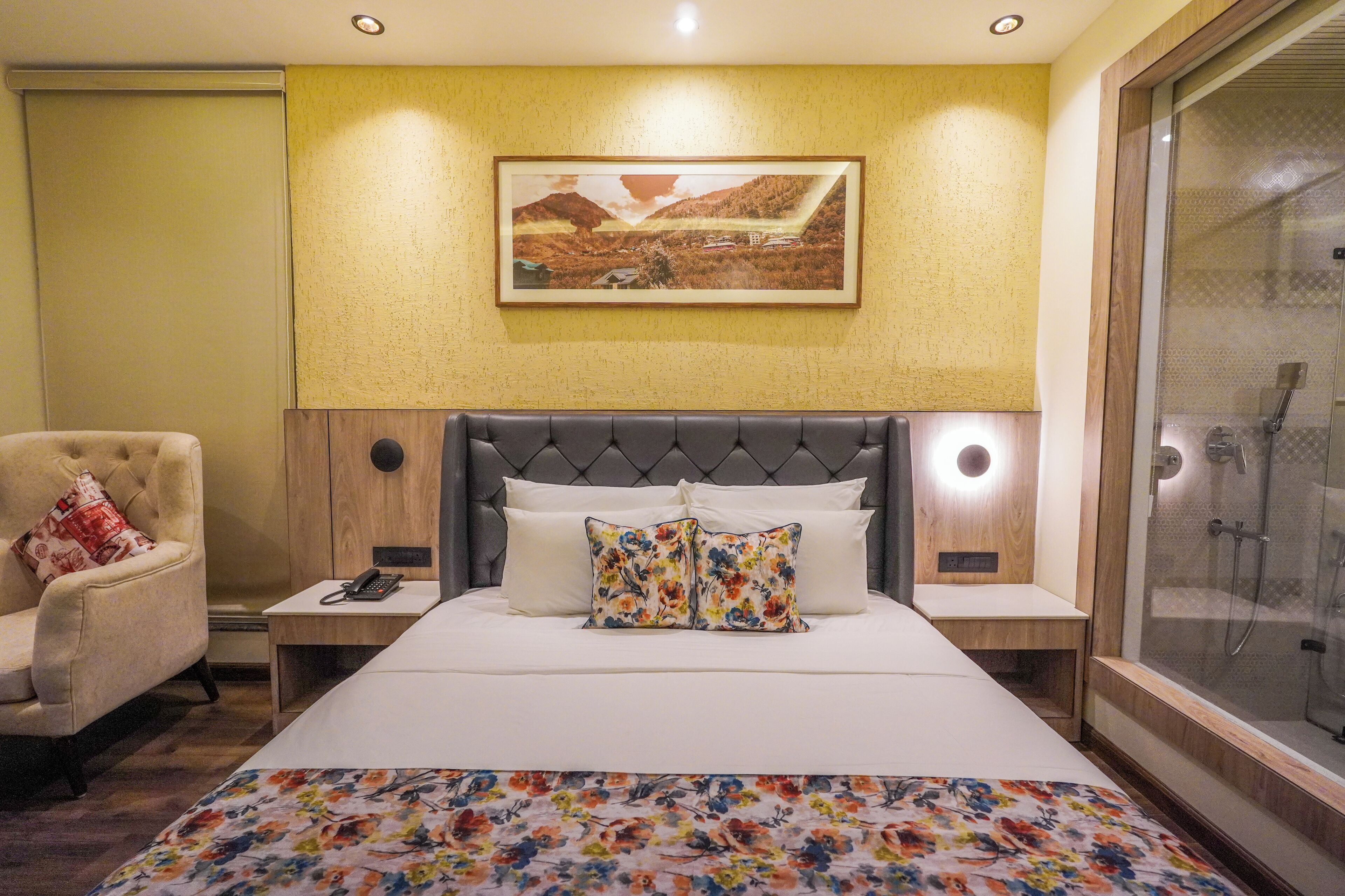 Habitación de hotel con una cama grande, una silla, una mesita de noche con teléfono y un cuadro en la pared, además de un box de ducha de vidrio.