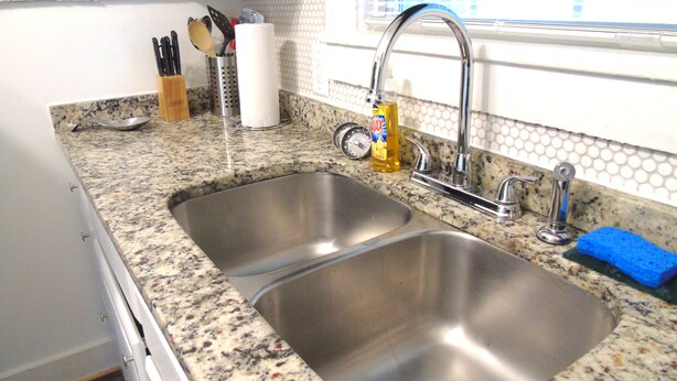 Una cocina con dos lavamanos de acero inoxidable, una ventana con persianas y una encimera con diversos utensilios de cocina.