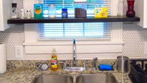 Una cocina con un lavamanos doble, una ventana con persianas y estantes con diversos utensilios de cocina.