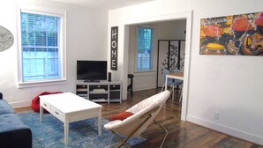 Una sala de estar con televisor, un sofá blanco, un sillón de peluche rojo, un tapete azul y un comedor con mesa y sillas.