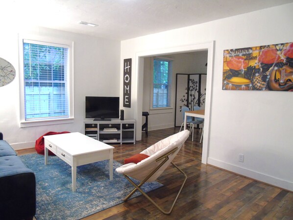 Un salon avec un téléviseur, un canapé blanc, un fauteuil poire rouge, un tapis bleu et un coin repas avec une table et des chaises.