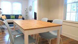 Un comedor con mesa de madera y cuatro sillas, un sofá con cojines euna lámpara de pie.