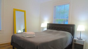 Una cama bien tendida con colcha gris y una toalla blanca doblada, una mesita de noche con lámpara, un espejo grande con marco amarillo y una ventana con persianas.