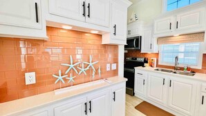 A kitchen with white cabinets, a brick backsplash, and a window with blinds. - Vacation Cottage North Myrtle Beach (North Myrtle Beach)