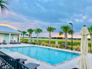A rectangular swimming pool with a clear blue water, surrounded by a concrete deck. There are lounge chairs and a covered umbrella on the deck. Palm trees are visible in the background. - Vacation Cottage North Myrtle Beach (North Myrtle Beach)