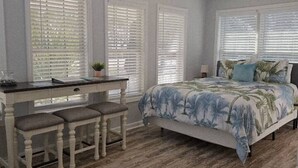 A bedroom with a bed, a desk with two chairs, a ceiling fan, and large windows with blinds. - Drift Away with Inlet Vibes in Murrells Inlet at a Peaceful Studio (Murrells Inlet)