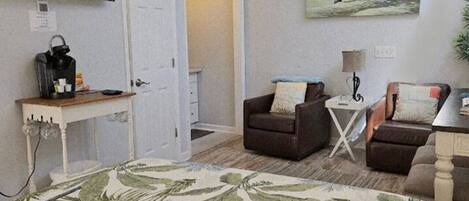 A bedroom with a bed, two armchairs, a desk with a coffee maker, a TV, and a painting of a beach scene.