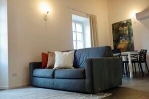 A modern living room with a grey sofa, a dining table, and a painting on the wall.