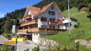 A two-story building with balconies, a stone staircase, and a view of a forested hillside. - Hotel Sterne - Economy Triple (Beatenberg)