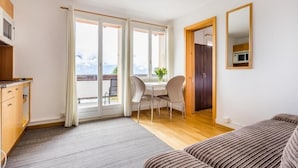 A modern room with a wooden floor, a bed, a dining table with chairs, a kitchen area with a microwave, and a sliding glass door leading to a balcony with a view. - Hotel Sterne - Economy Triple (Beatenberg)