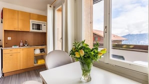 A small kitchenette with wooden cabinets and a microwave, a dining table with a vase of flowers, and a view of a mountain through a large window. - Hotel Sterne - Economy Triple (Beatenberg)