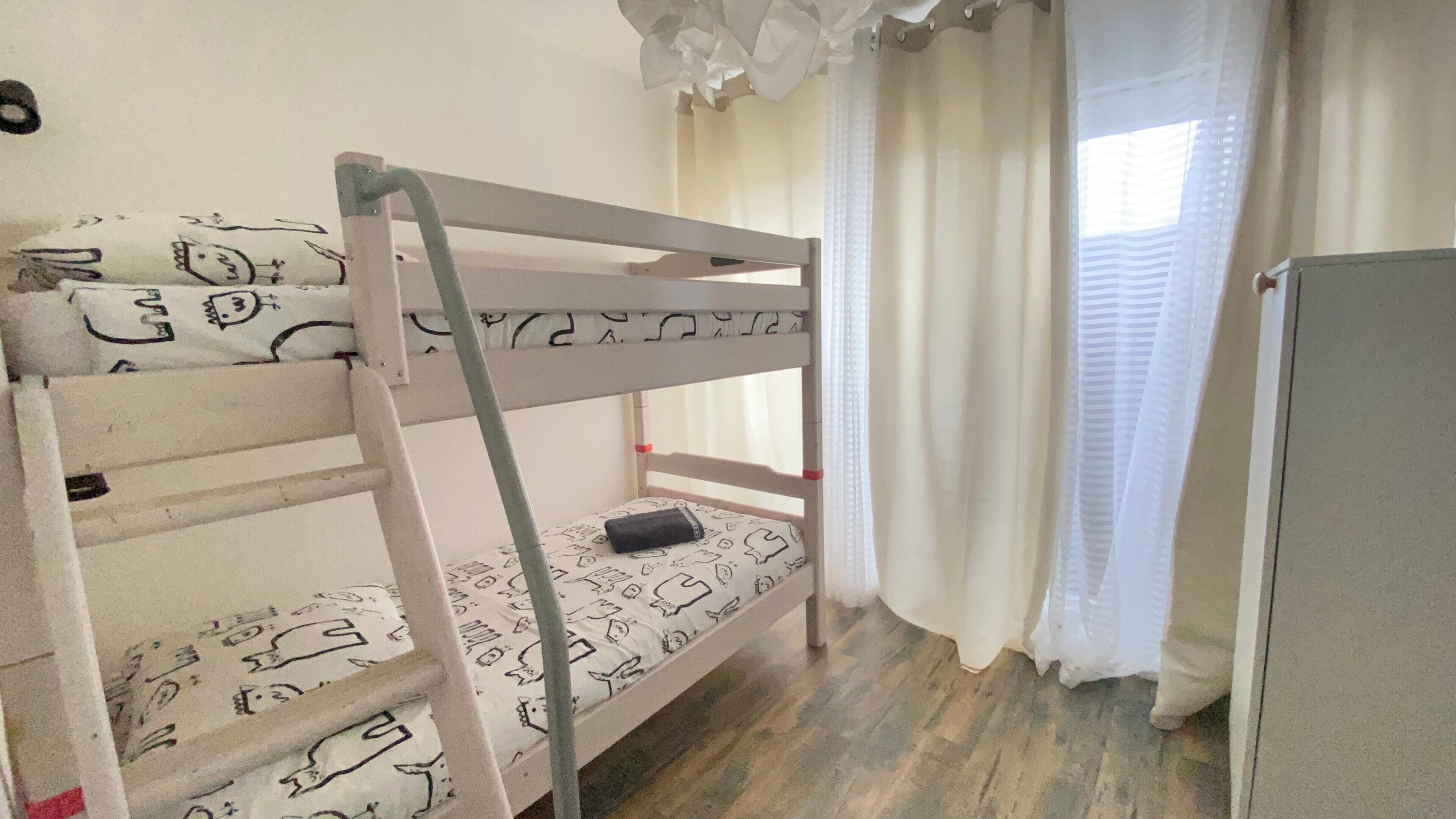 Una cama individual superpuesta con escalera, una mesita de noche pequeña y una ventana con cortinas.
