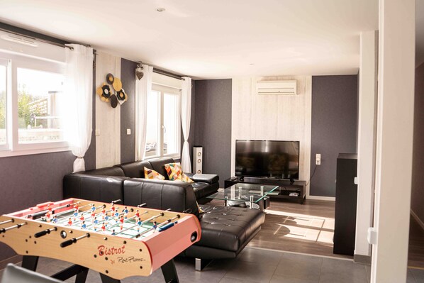 A living room with a foosball table, a black leather sofa, a glass coffee table, and a flat-screen TV.