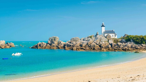A coastal scene with a lighthouse, rocky shoreline, clear blue water, and a sandy beach.