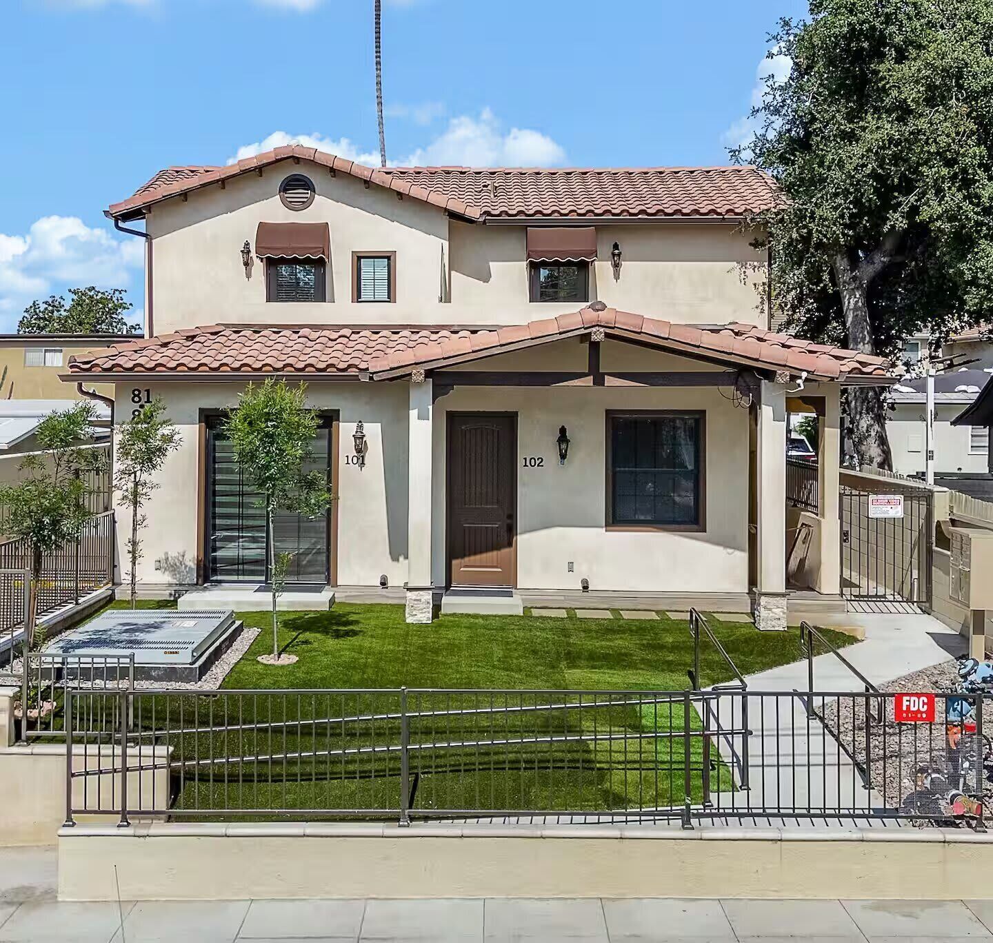 A two-story house with a tiled roof, numbered 101 and 102, featuring a front porch, a small lawn, and a gated driveway.