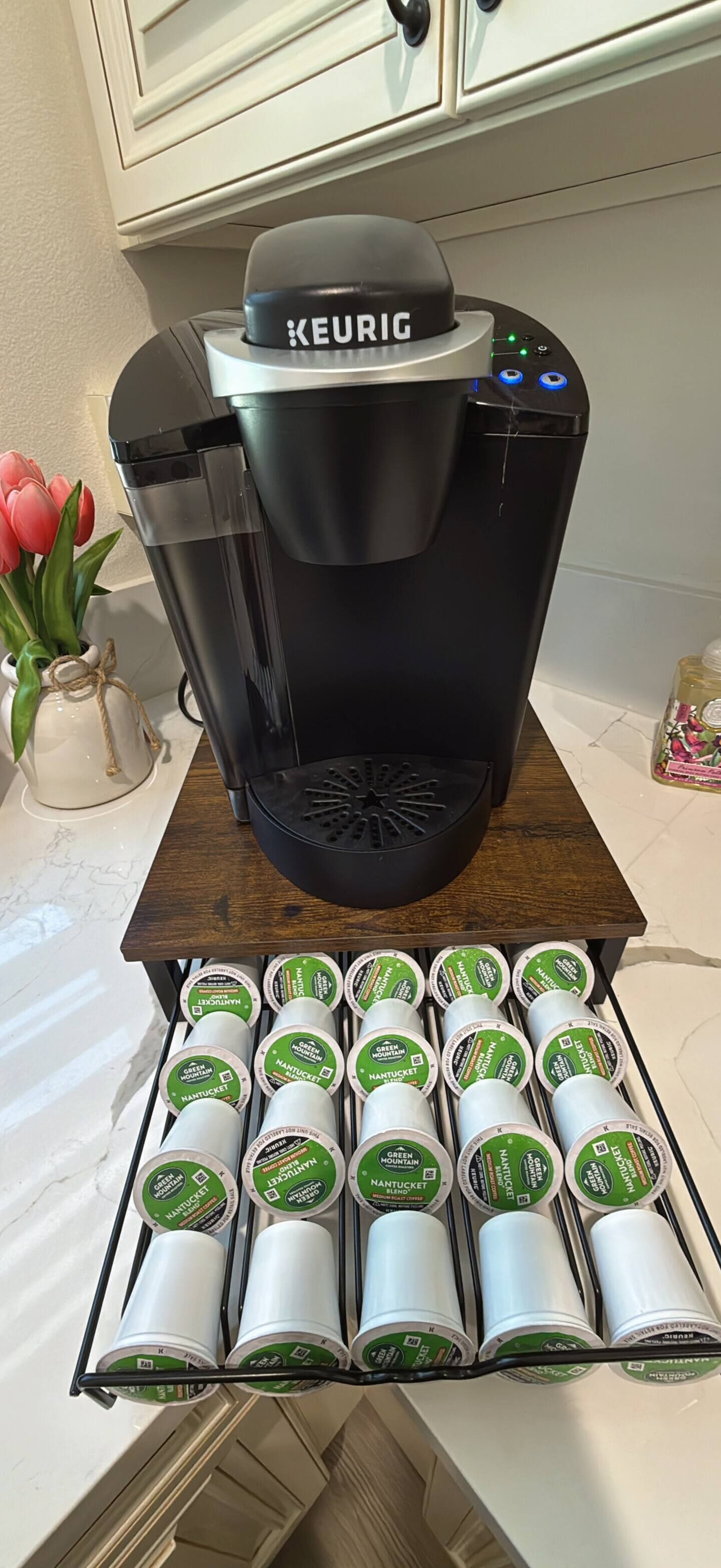 Eine Keurig-Kaffeemaschine auf einem Holztisch, darunter eine Schale mit Nannoticket-Kaffeekapseln.