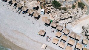 An aerial view of a beach with rows of sun loungers and umbrellas, rocky terrain, and a clear view of the sea. - Intimate Studio w Garden and Inviting Shared Pool (Samos)