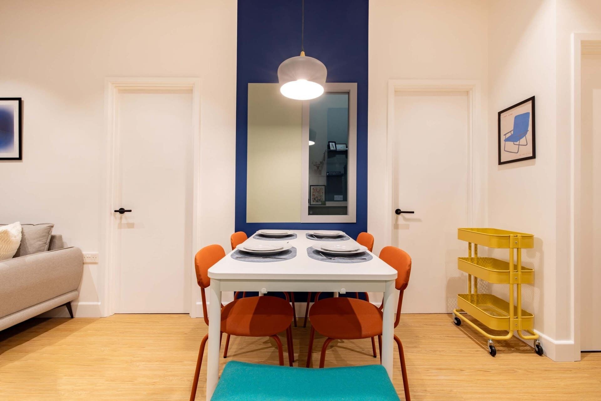 Un coin repas avec une table blanche, des chaises orange, un mur bleu et un chariot jaune.