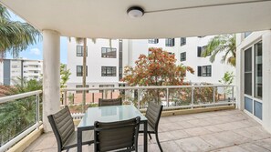 Een balkon met een glazen tafel en vier stoelen, met uitzicht op een gebouw met veel ramen en een boom met rode bloemen.