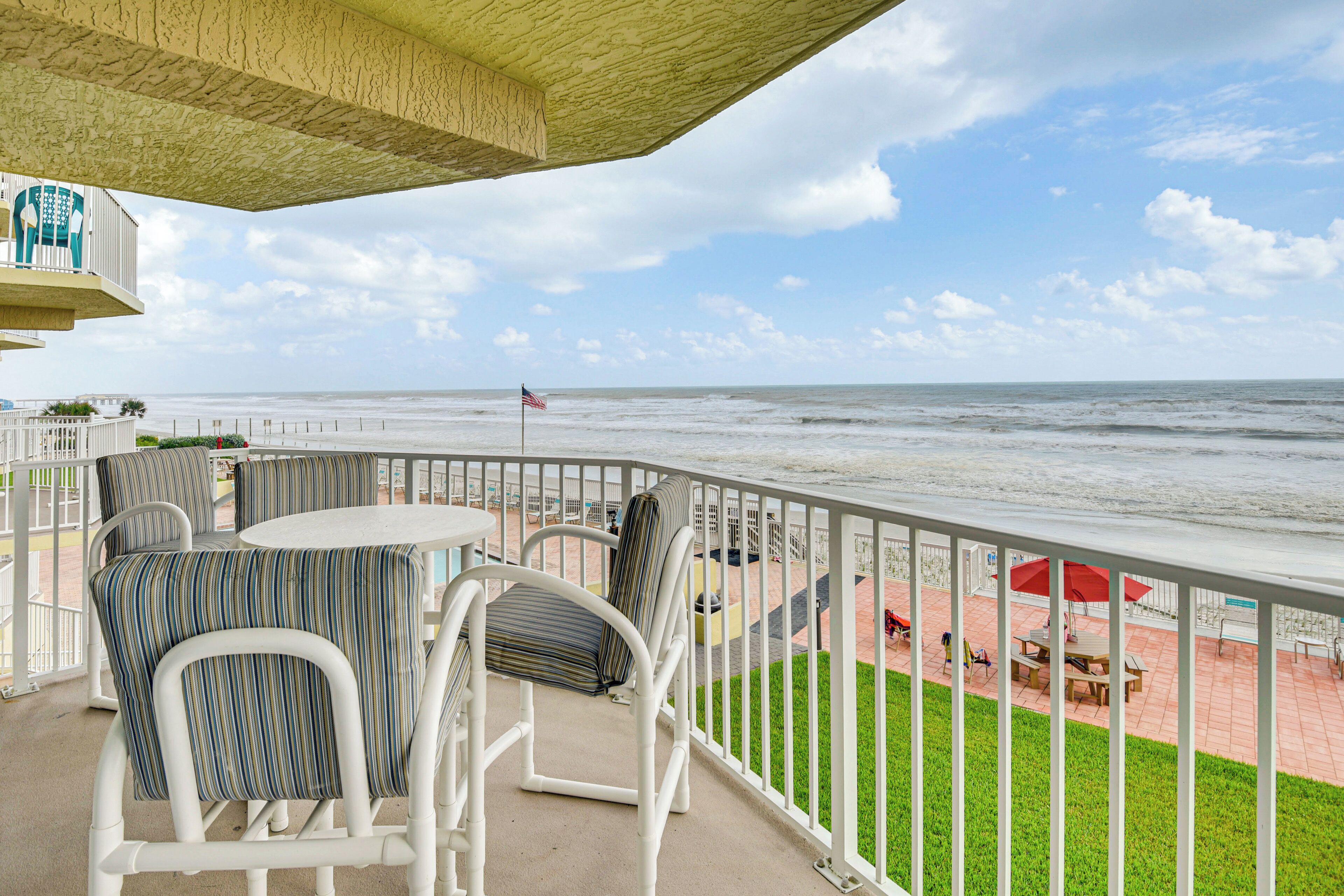 Ein Balkon mit weißen Stühlen und einem Tisch, der Blick auf einen Strand mit einer Flagge bietet.