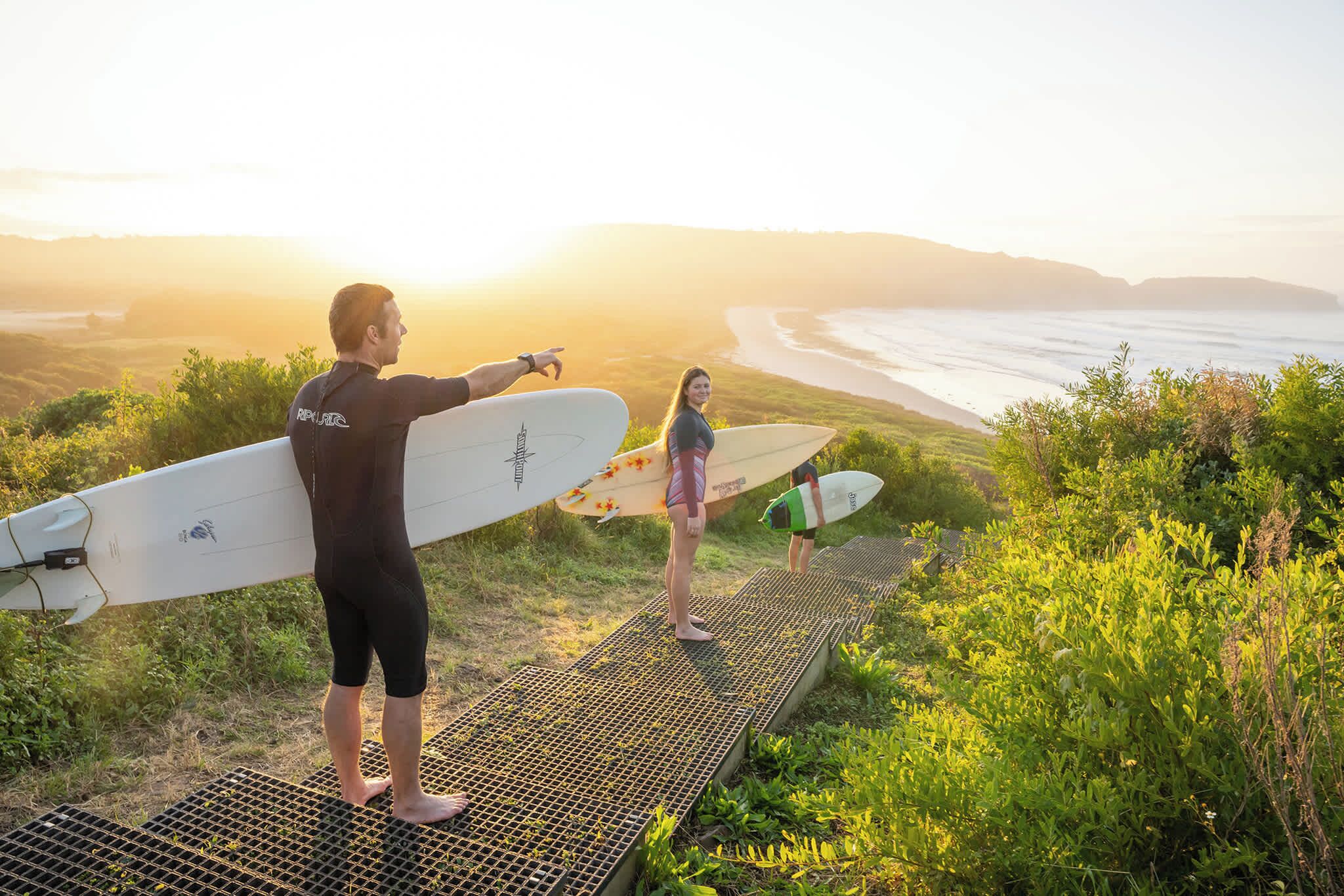 Eine Person hält ein Surfbrett auf einer Plattform mit Blick auf den Ozean und die dahinterliegenden Hügel.