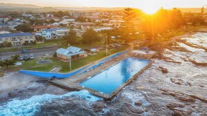 A swimming pool adjacent to a rocky shoreline with a view of a residential area and a building with a solar panel. - The Nook - Lake Illawarra (Lake Illawarra)